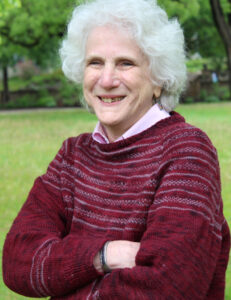 A white woman with white hair wearing a burgundy sweater crossing her arms and smiling in front of a greenery background