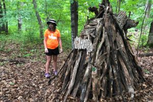 Teen female standing in woods next to shelter made of fallen trees.