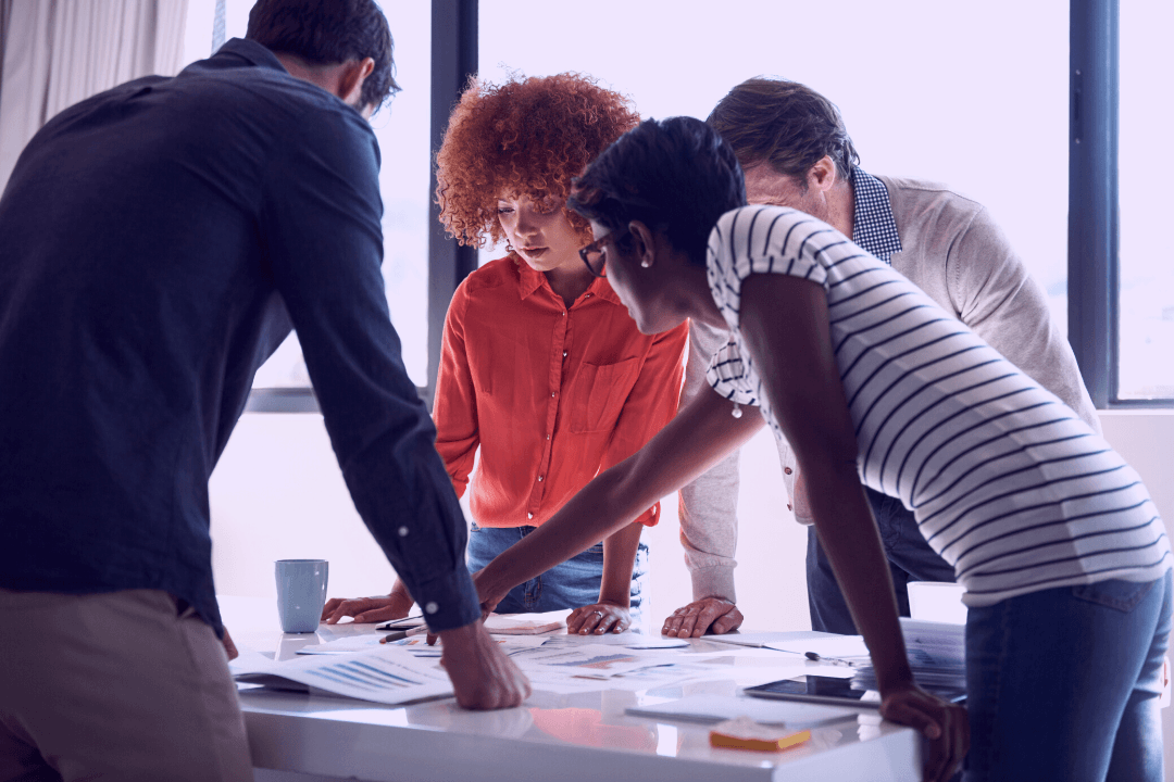 A group of diverse young adults gathers around a table. One of them points something out.