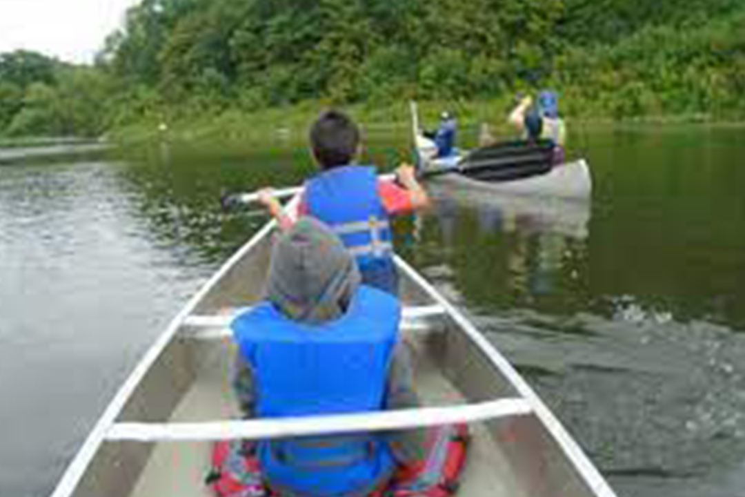 Image of two kids in a canoe wearing blue life jackets and paddling on the water