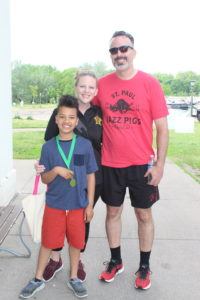 Photo of family featuring a mom in a black shirt, a dad in a red shirt with black shorts and wearing sunglasses, and a young boy with red shorts and a blue shirt, proudly showing his walk medal