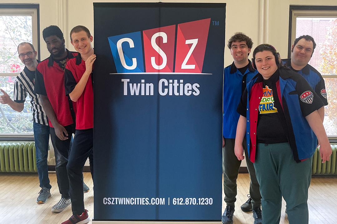 A group of young adults in matching bowling shirts poses in front of a screen with the CsZ logo