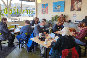 A coffee shop with autistic adults seated at tables. Some are talking, some are working on projects, and others are just hanging out.