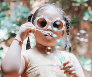 A girl with amazing flower shaped sunglasses and cute pigtails blows bubbles towards the camera