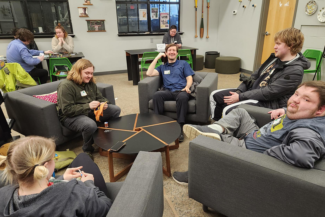 A group of adults sits in a circle of comfortable chairs at a coffee shop and talks animatedly