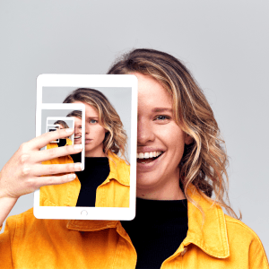 A woman looks at the camera smiling. She's holding a photo of herself looking neutral. In that photo she's holding another photo with a different emotion.