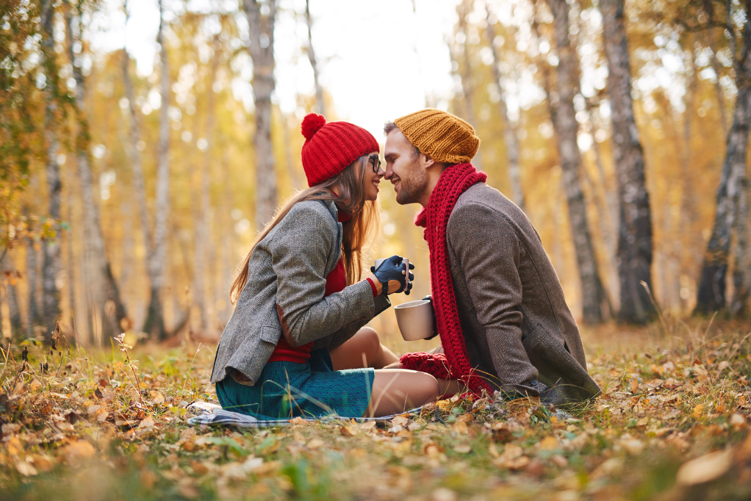 A man and a woman sit on a blanket in the woods, nose to nose, with mugs in their hands