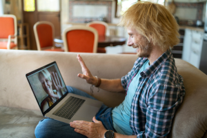 A man sits on his couch with his laptop open. There is a video chat happening and he's waving to the woman he's chatting with