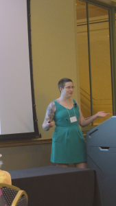 A nonbinary individual stands at the front of the room giving a presentation and gesturing with their hands