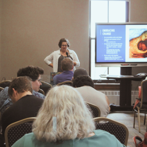 A woman stands next to a slide presentation, speaking into a microphone