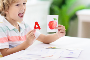 A young boy holds up two cards. One shows an apple and the other has the letter A.