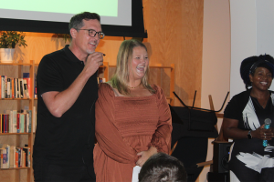 AuSM's Executive Director Ellie Wilson stands next to a man speaking into a microphone in front of a crowd at Brother Justus. They're both smiling widely.
