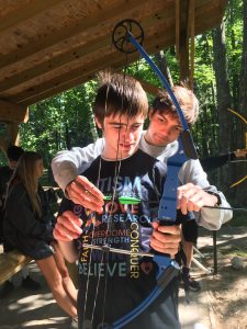 A camp counselor stands behind his camper and helps to nock an arrow to a bow