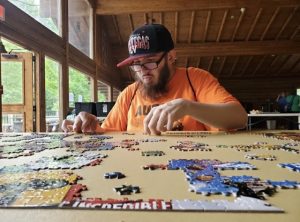 An adult camper sits at a table with a partially finished puzzle. He intently looks for the space for his next piece.