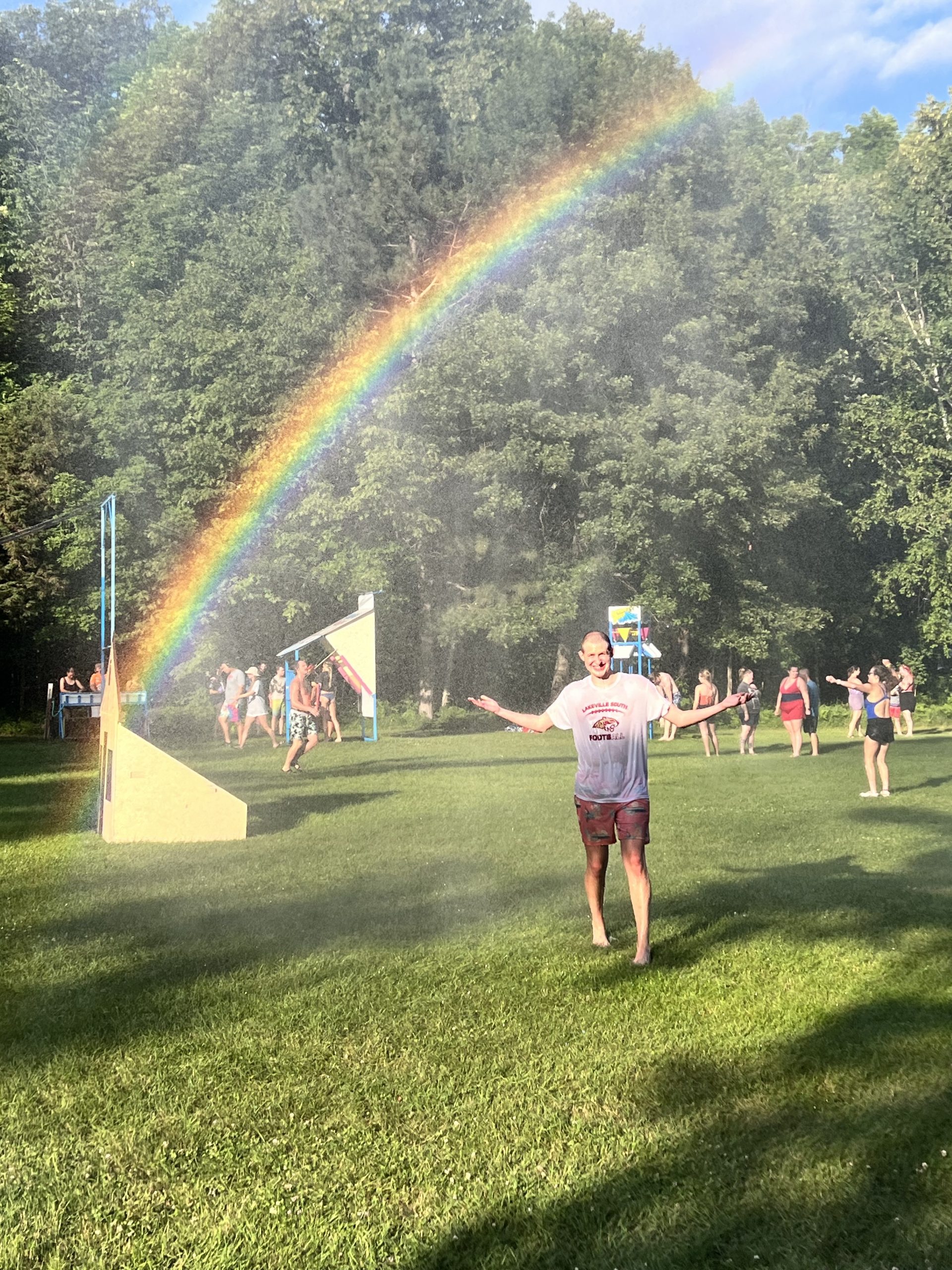 A camper, a young man, stands in a field underneath a spray of water, soaking it up. There's a beautiful rainbow over his head.