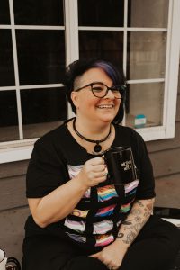 A nonbinary individual holding a coffee cup and grinning widely