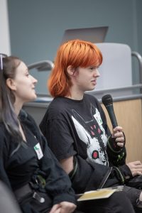 A young individual holds a microphone. They have bright red hair and wear a dark shirt.