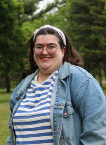 A white nonbinary person with brown hair wearing a white bandana in their hair, a light wash jean jacket, and a white and light blue striped shirt smiling in front of a greenery background
