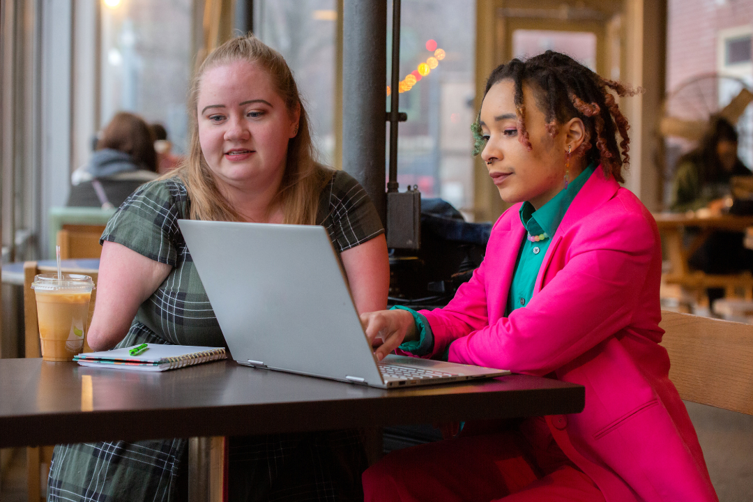 Two individuals sitting at a table in a cozy cafe, engaged in collaborative work on a laptop. The person on the left has light skin, long blonde hair, and wears a green plaid dress. The person on the right has medium skin, styled braids, and is dressed in a bright pink suit with a teal shirt. A notebook, pen, and iced coffee sit on the table, adding to the productive and friendly atmosphere.