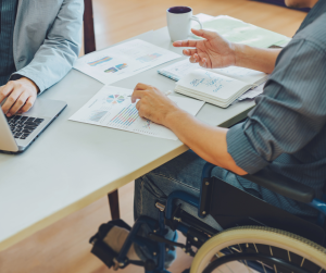 Two individuals collaborating at a desk with charts and graphs spread out. One person is using a laptop, and the other, seated in a wheelchair, gestures while discussing ideas. A notebook with handwritten notes and a coffee mug are visible on the table.