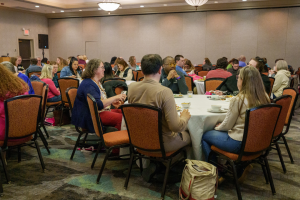 A large group of people seated around round tables in a conference room. The participants are engaged in conversations, with plates and cups on the tables. The room has a neutral-colored interior with warm lighting and a carpeted floor.