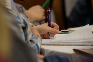 A close-up of a hand writing in a notebook with a purple pen. The background shows blurred details of other attendees holding pens and notebooks, suggesting a workshop or seminar.