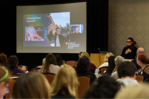 A speaker presenting to an audience in a conference room. A slide is displayed on a large screen, featuring text and images under the title "A Growing Global Movement." The audience members are seated and focused on the presentation. A sign language interpreter stands to the right of the screen, gesturing.