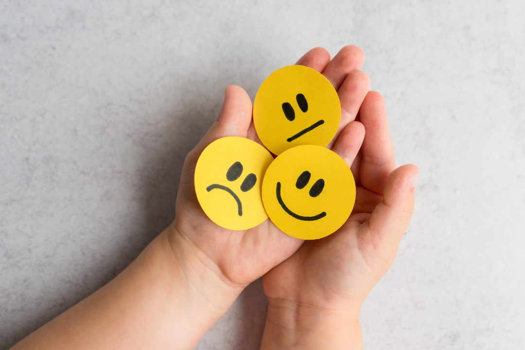 A pair of hands holding three yellow circular cutouts with different facial expressions: a sad face, a neutral face, and a smiling face, symbolizing a range of emotions.