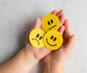 A pair of hands holding three yellow circular cutouts with different facial expressions: a sad face, a neutral face, and a smiling face, symbolizing a range of emotions.