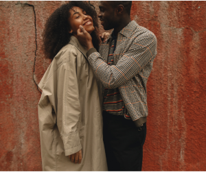 A man and a woman stand in front of a red wall. They are in a half hug and the man is brushing his fingers over the woman's cheek while she smiles at him.