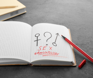 "An open notebook with grid paper showing symbols for male, female, and a question mark, alongside the words 'SEX education' written in red ink. A red pen is placed on the page, with a capped red marker nearby on a gray table."