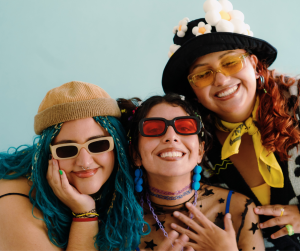 Three friends smiling and posing together in colorful, fun outfits and accessories. One person has blue braided hair, wearing a beige beanie and rectangular cream-colored sunglasses. Another has short dark hair, red sunglasses, and vibrant earrings. The third has curly red hair, a black hat with flower details, yellow-tinted glasses, and a yellow scarf. The background is a solid light blue.