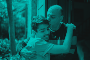 A father gives his son a side hug and kisses him on the forehead. The image is tinted teal.
