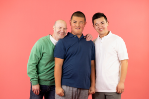 Three people stand closely together in front of a solid pink background, smiling at the camera. A man in a green sweater is on the left, a young man in a navy blue polo shirt is in the center, and another young man in a white polo shirt is on the right with his arm around the person in the center.
