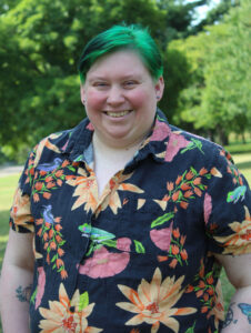 A white trans man with neon green hair wearing a navy blue floral button up shirt smiling in front of a greenery background