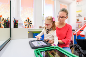 A young girl with glasses and a pink bow in her hair uses a tablet with the assistance of a woman, likely a caregiver or therapist, who is seated behind her. They are in a brightly lit room with colorful decorations, art supplies, and a wheelchair visible in the background. The scene suggests a supportive, therapeutic, or educational environment focused on assistive technology.
