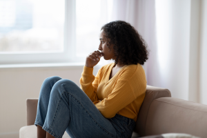 A young woman with curly hair sits on a couch with her knees drawn up to her chest, looking out the window with a thoughtful, concerned expression. She is wearing a mustard yellow sweater and blue jeans. Soft daylight filters through the curtains behind her.
