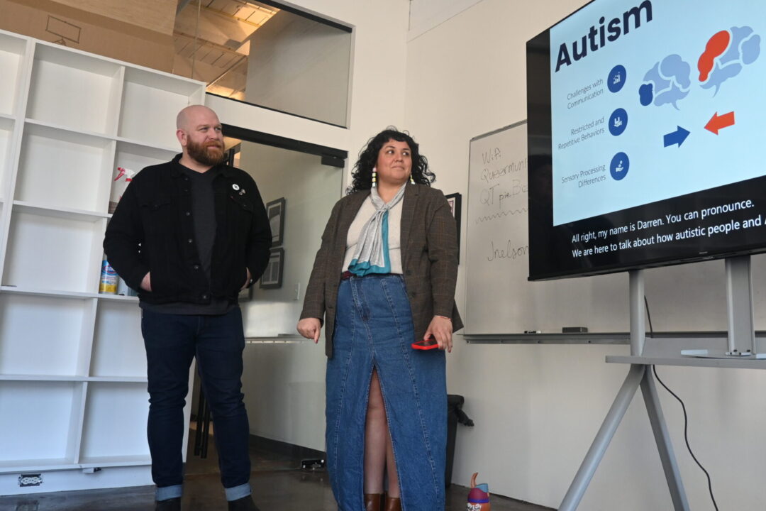 A white man wearing a black coat and black jeans standing next to a woman of color wearing a green khaki coat, a jean skirt, and a light blue scarf looking at a tv screen that reads 