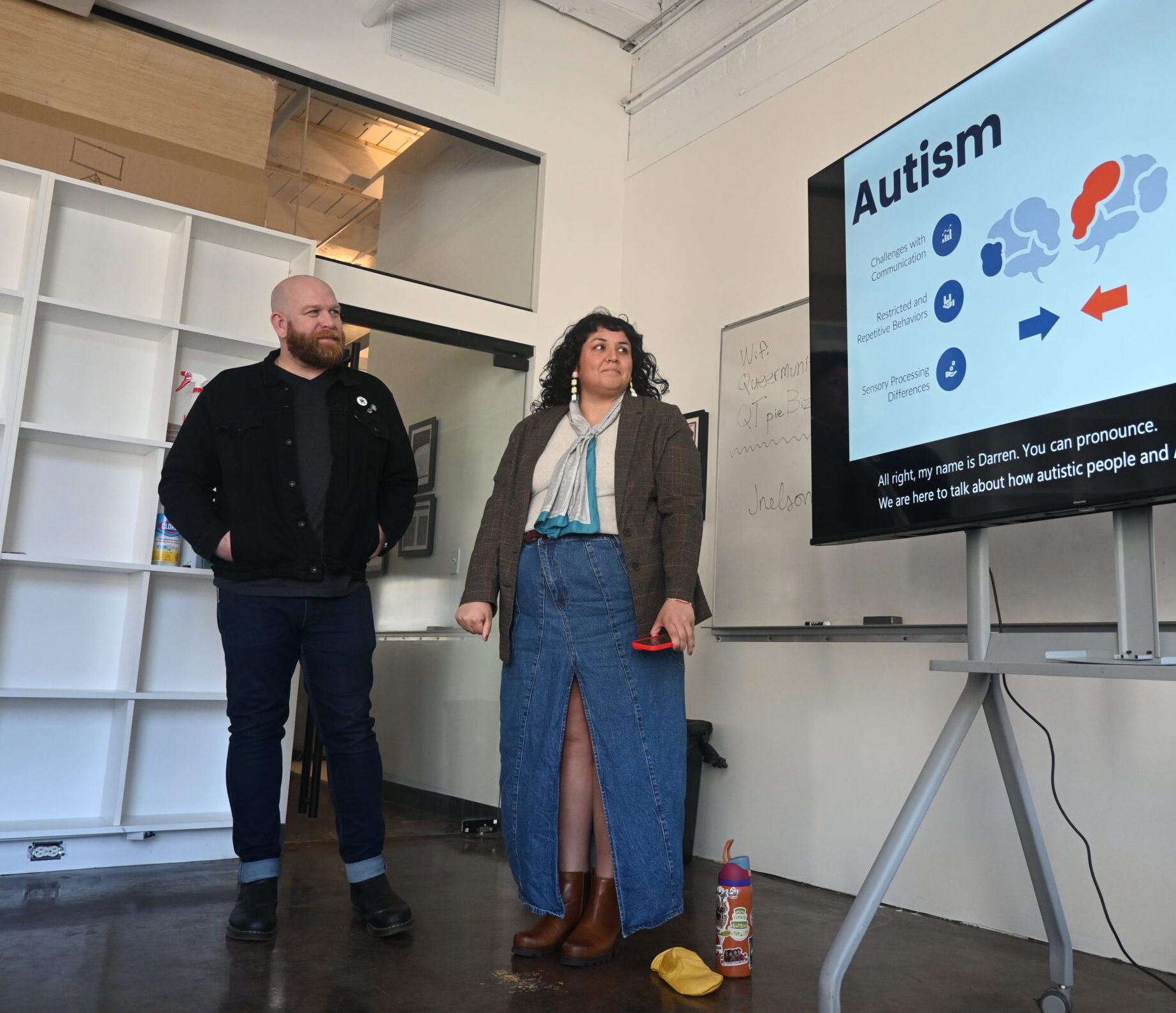 A white man wearing a black coat and black jeans standing next to a woman of color wearing a green khaki coat, a jean skirt, and a light blue scarf looking at a tv screen that reads "autism: challenges with communication, restricted and repetitive behaviors, sensory processing differences."