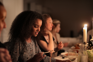 A group of women sit at a warmly lit dinner table, engaged in quiet conversation and eating. The focus is on a young woman with curly hair and a floral blouse, looking down at her plate with a look of disgust while holding a fork. A candle glows in the foreground, creating a soft, intimate atmosphere.