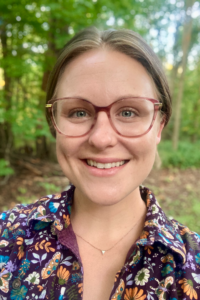 A person smiling outdoors in a wooded area. They have light skin, short brown hair parted in the middle, and are wearing large round glasses, a small triangular necklace, and a purple shirt with a colorful floral pattern.