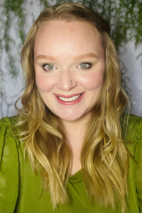 A smiling woman with long wavy blonde hair wearing a green blouse. They are in front of a softly blurred background with green leafy plants.