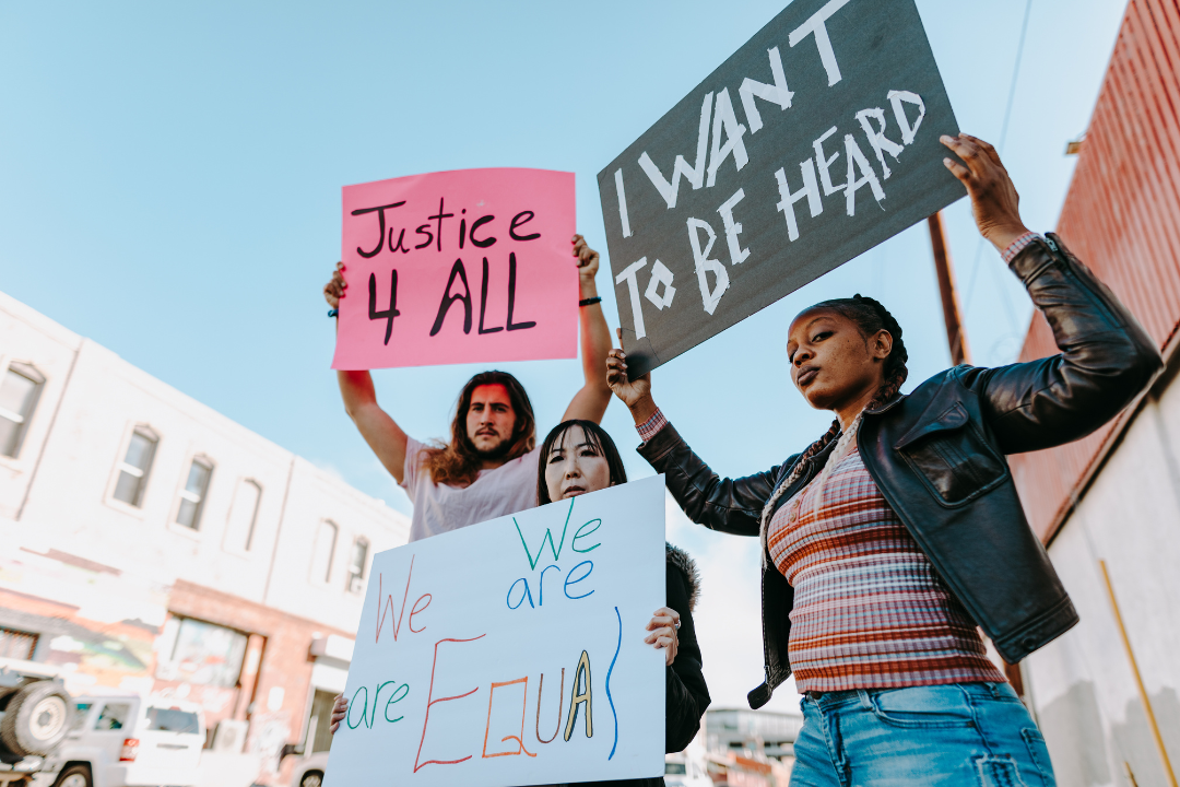 Three people at a protest hold signs in the air. One sign reads “Justice 4 All,” another reads “I Want to Be Heard,” and the third says “We are Equal.”