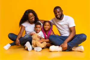 A Black family of four sits together smiling against a bright yellow background. The mother and father sit on either side of their two children, who are holding a teddy bear.