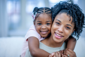 A young Black girl with braids and beads smiles as she hugs her mother from behind. Both are smiling warmly in a close-up portrait.