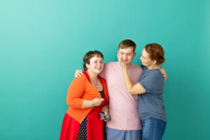 Three people stand close together in front of a teal background, smiling and embracing. A young person in a light pink shirt stands in the middle, with a person in a red cardigan on the left and a person in a blue shirt on the right. The person on the right gently touches the face of the person in the middle, creating a warm, affectionate moment.