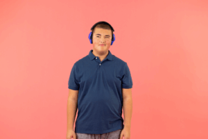 A young person stands against a solid coral-pink background, wearing a navy blue polo shirt and gray shorts. They have short hair and are wearing bright blue over-ear noise-canceling headphones. They face the camera with a calm, relaxed expression and their arms resting at their sides.