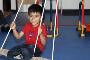 A smiling child sits on a rope swing inside an indoor gym, holding the ropes with both hands. The child wears a red T-shirt and jeans, with sensory play equipment visible in the background on a blue padded floor.