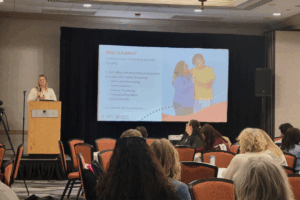 A presenter stands at a podium in a hotel conference room while a slide titled “What is Autism?” is projected behind them. The slide explains autism as a brain-based developmental disability and lists areas it can impact, such as communication, sensory processing, and emotional regulation. Audience members sit at round tables facing the stage.