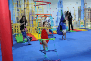 Children play in a colorful indoor sensory gym with padded flooring, climbing structures, rope ladders, swings, and a small climbing wall. One child climbs a rope ladder in the foreground while others explore equipment nearby, with adults supporting children in the background.